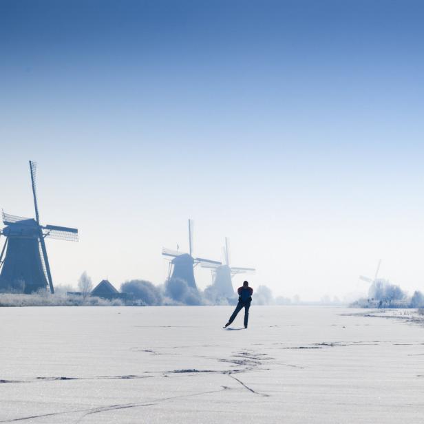 Eine Person läuft Schlittschuh auf einem zugefrorenen See mit Windmühlen im Hintergrund.