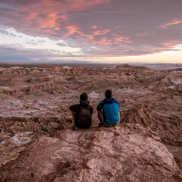 Zwei Personen sitzen auf einem Felsen und blicken auf die Wüstenlandschaft bei Sonnenuntergang.