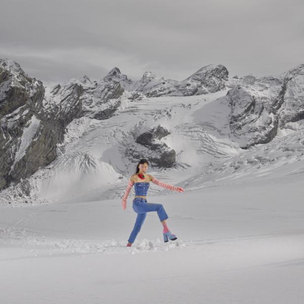 Eine Frau in Jeanskleidung watet durch den Schnee vor einer Bergkulisse.