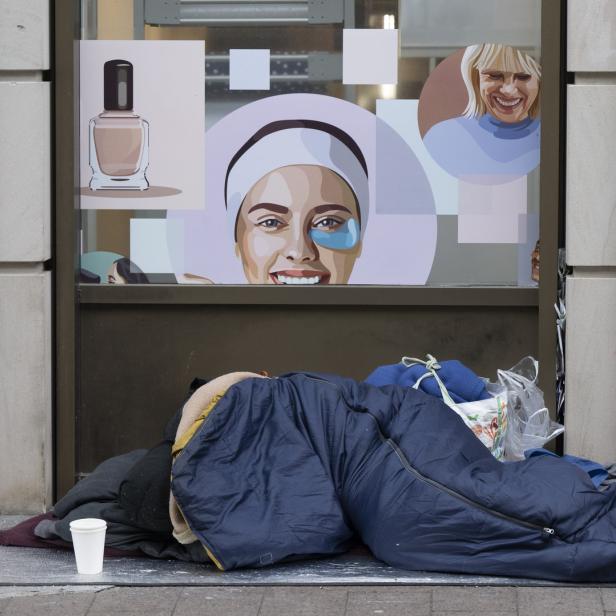 Eine Person liegt in einem Schlafsack vor einem Geschäft mit Kosmetikwerbung.