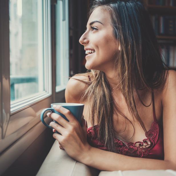 Eine Frau lächelt, während sie mit einer Tasse Kaffee am Fenster sitzt.