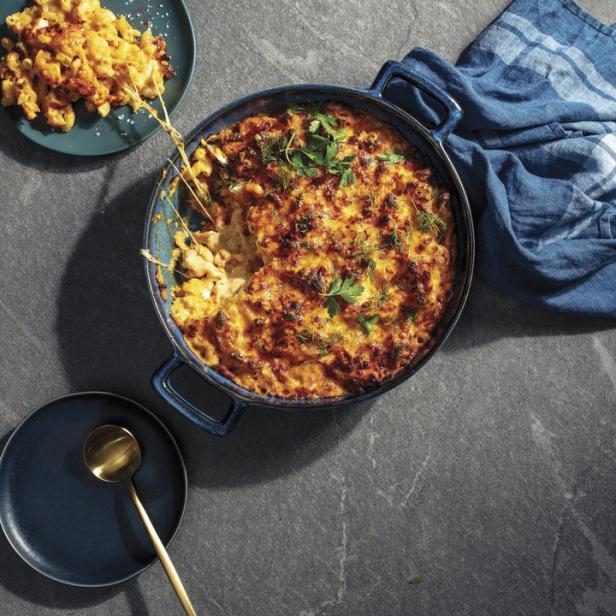 Ein Auflauf mit Mac and Cheese wird mit einem goldenen Löffel und einem blauen Geschirrtuch präsentiert.
