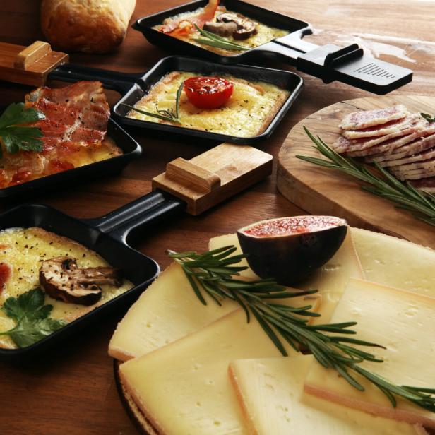 Mehrere Raclettepfännchen mit geschmolzenem Käse, Gemüse und Speck, dazu Käse- und Wurstscheiben mit Rosmarin.