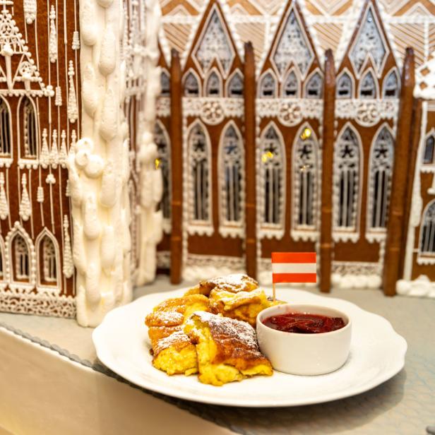 Ein Teller Kaiserschmarrn mit Zwetschgenröster vor einem Lebkuchenhaus mit österreichischer Flagge.
