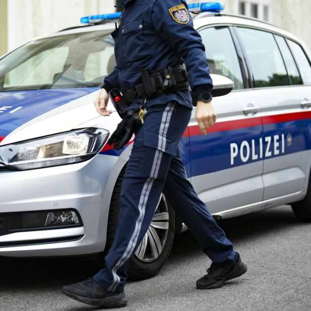 Ein Polizist in Uniform geht an einem silbernen Polizeiauto mit blauen und roten Streifen vorbei.