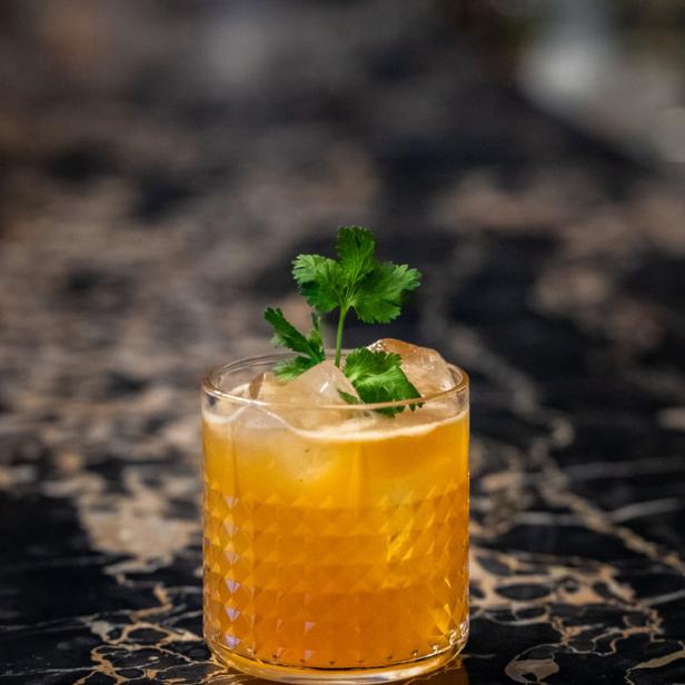 Ein Cocktail mit Eiswürfeln und einem Zweig Koriander auf einem dunklen Tisch.