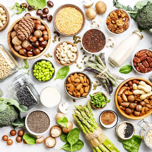 Verschiedene vegane Lebensmittel wie Nüsse, Samen, Hülsenfrüchte, Sojabohnen, Spargel, Champignons, Spinat und Brokkoli auf einem Tisch.