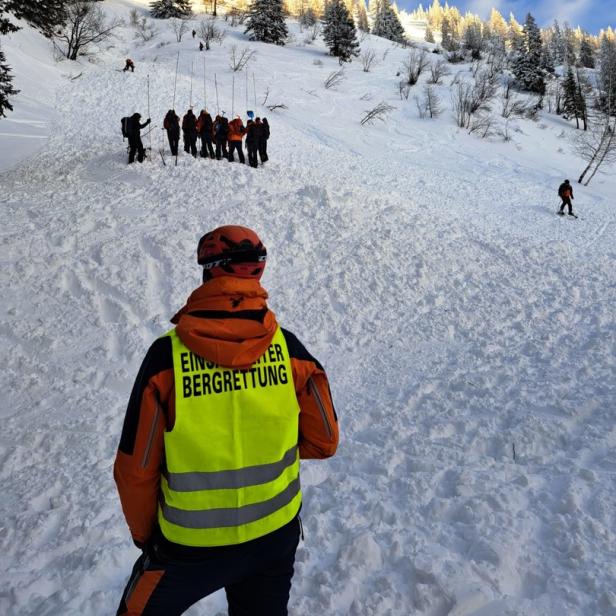 Ein Mitglied der Bergrettung steht vor einem schneebedeckten Hang mit Suchmannschaft.