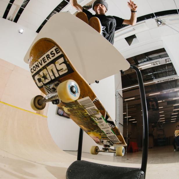 Ein Skateboard mit Converse-Logo steht auf einer Halterung, ein Skater im Hintergrund mit erhobenen Armen.
