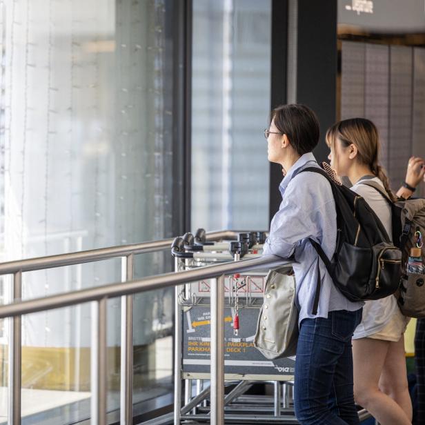 Zwei Frauen mit Rucksäcken stehen an einem Flughafenfenster und schauen hinaus.