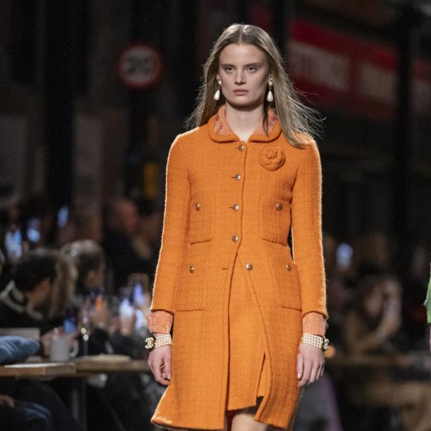 Ein Model präsentiert einen orangefarbenen Tweed-Mantel mit passendem Rock auf dem Laufsteg.