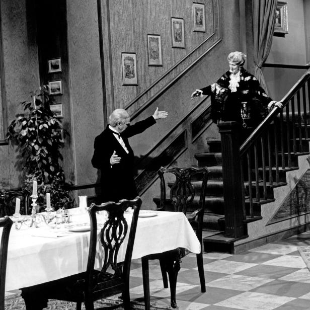 Ein Mann im Smoking begrüßt eine Frau auf einer Treppe in einem eleganten Haus.