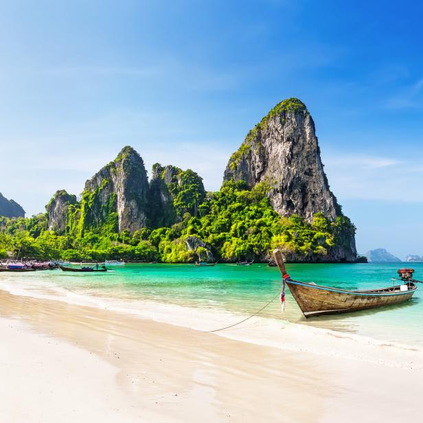 Ein traditionelles thailändisches Boot liegt an einem tropischen Strand mit Kalksteinfelsen im Hintergrund.