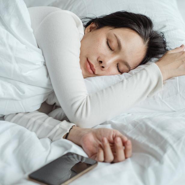 Eine Frau schläft in einem weißen Bett, neben ihr liegt ein Smartphone.