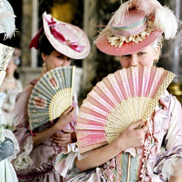 Frauen in historischen Kostümen und Hüten verdecken ihre Gesichter mit Fächern.