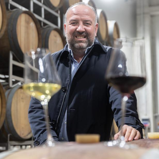 Ein lächelnder Mann steht vor Weinfässern mit zwei Gläsern Wein.