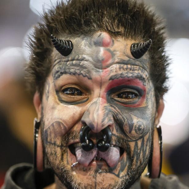 Ein Mann mit Gesichtstattoos, Hörnern auf der Stirn und Piercings in Nase und Lippen streckt die Zunge heraus.