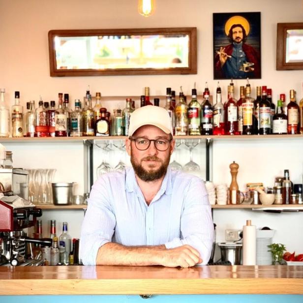 Ein Mann mit Bart und Kappe steht hinter einer Bar mit Spirituosenflaschen und Gläsern.