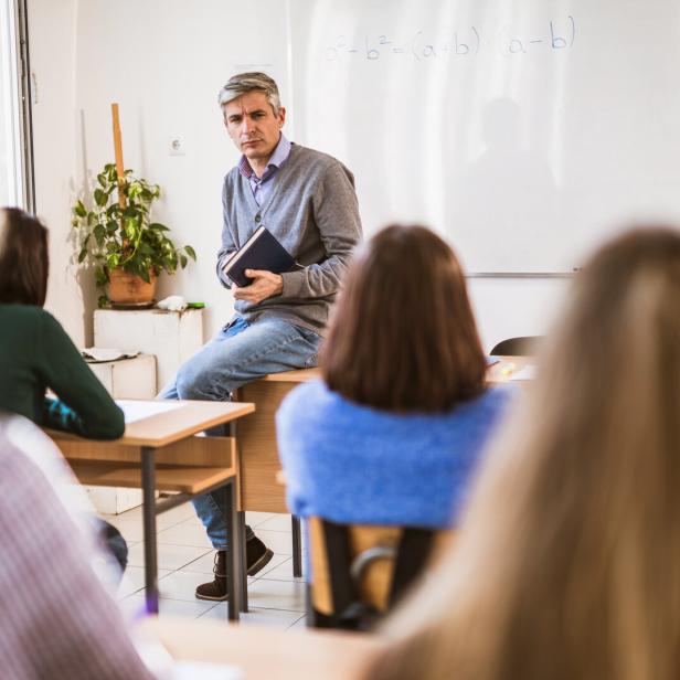 Ein Lehrer sitzt auf einem Tisch vor einer Klasse und hält ein Buch.