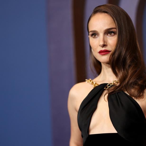 Natalie Portman in einem schwarzen Kleid mit goldenen Ketten vor einem blauen Hintergrund.
