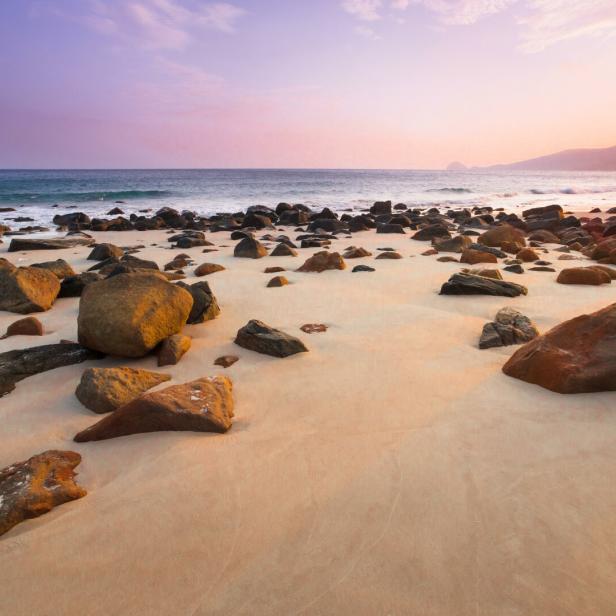 Ein sandiger Strand mit Felsen und einem Berg im Hintergrund bei Sonnenuntergang.