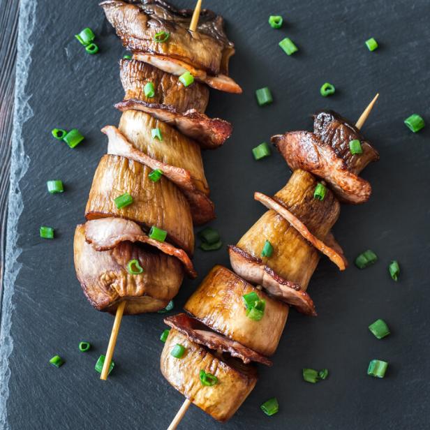 Zwei Spieße mit gegrillten Pilzen und Speck, garniert mit Schnittlauch, liegen auf einer Schieferplatte.
