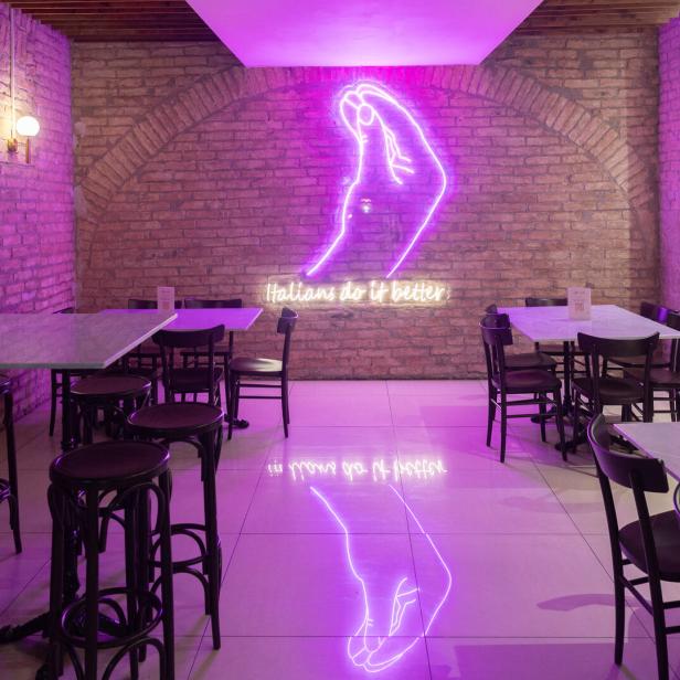 Ein Restaurant mit Tischen und Stühlen, beleuchtet mit violettem Neonlicht und dem Schriftzug „Italians do it better“.