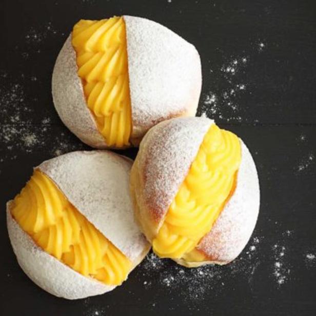 Drei mit gelber Creme gefüllte und mit Puderzucker bestäubte Krapfen liegen auf einem schwarzen Untergrund.