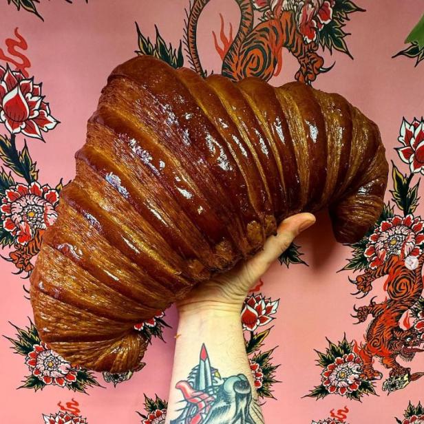 Eine Hand hält ein riesiges, glänzendes Croissant vor einem rosa Hintergrund mit Blumen- und Tigermuster.