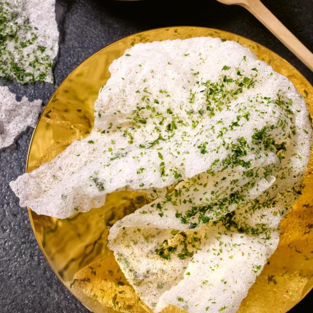 Ein goldener Teller mit Reiscrackern, garniert mit grünen Kräutern, neben einem Gericht mit rohem Fisch und Apfelstreifen.