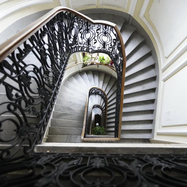 Blick nach unten durch ein Treppenhaus mit geschwungenem, verziertem Geländer und Pflanzen im unteren Bereich.