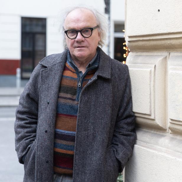 Ein Mann mit Brille und grauem Haar lehnt an einer hellen Wand.