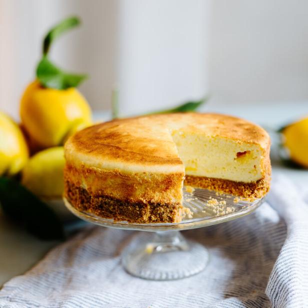 Ein angeschnittener Zitronenkäsekuchen auf einem Glasständer mit Zitronen im Hintergrund.