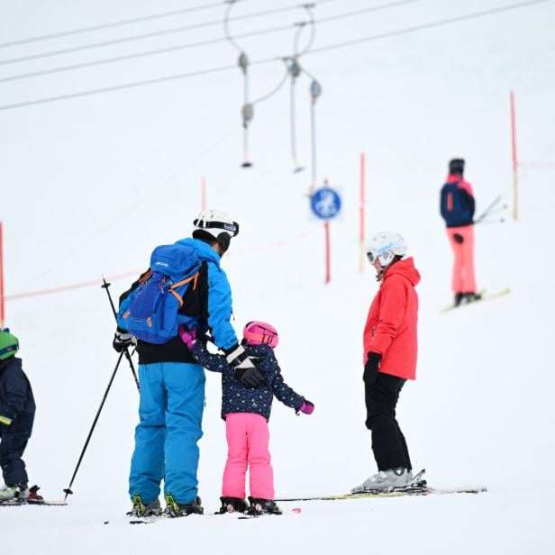 Mehrere Menschen, darunter Kinder und Erwachsene, stehen mit Skiausrüstung auf einer verschneiten Piste.