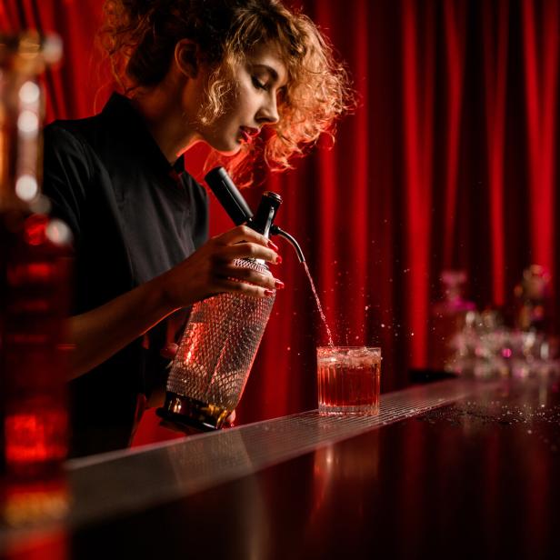 Eine Barkeeperin mixt einen Cocktail vor einem roten Vorhang.
