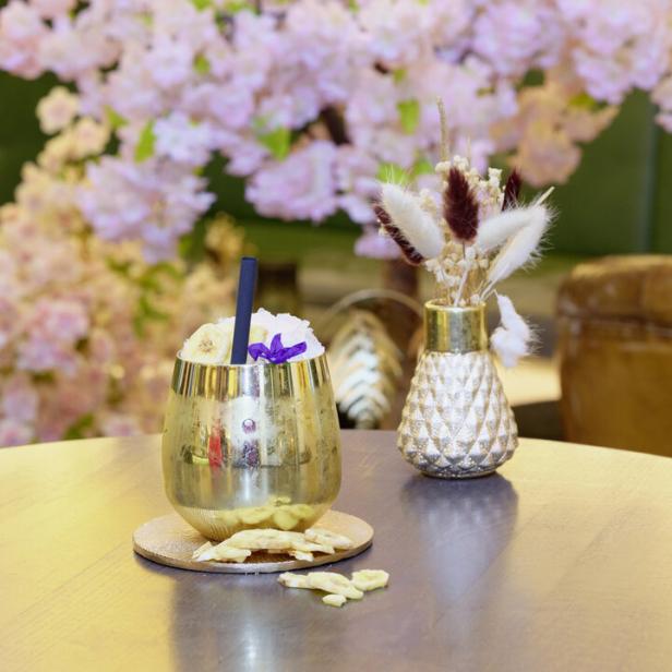 Ein goldener Cocktail mit Bananenchips und einer blauen Blüte vor einem rosa blühenden Baum.