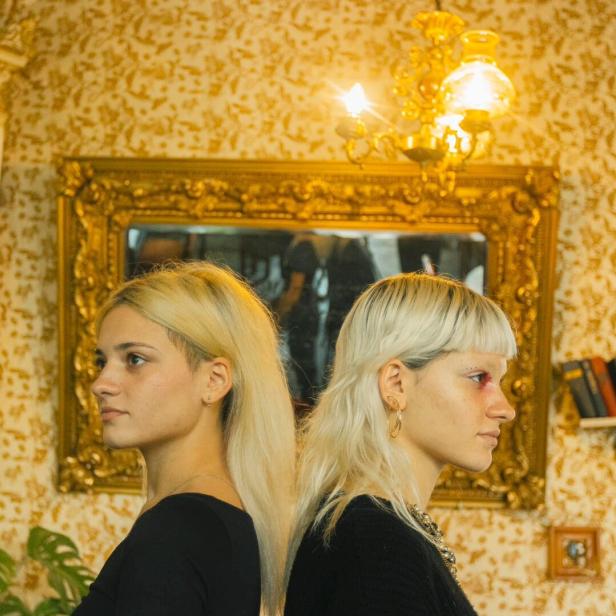 Zwei junge Frauen mit blonden Haaren stehen Rücken an Rücken vor einer goldenen Wand.