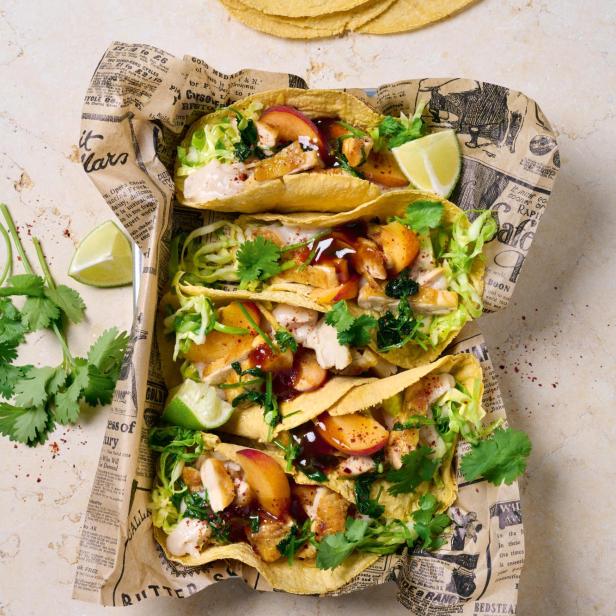 Drei Tacos mit Hähnchen, Salat, Limetten und einer dunklen Soße auf Zeitungspapier angerichtet.