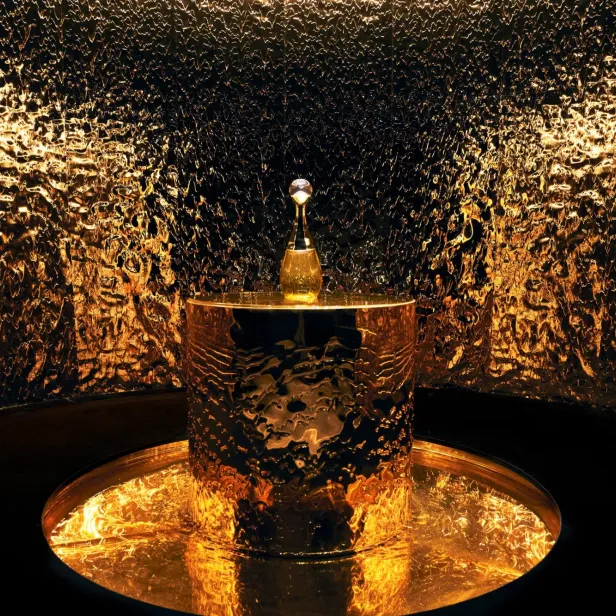 Ein goldener Parfümflakon steht auf einem goldenen Sockel vor einer goldenen Wand.