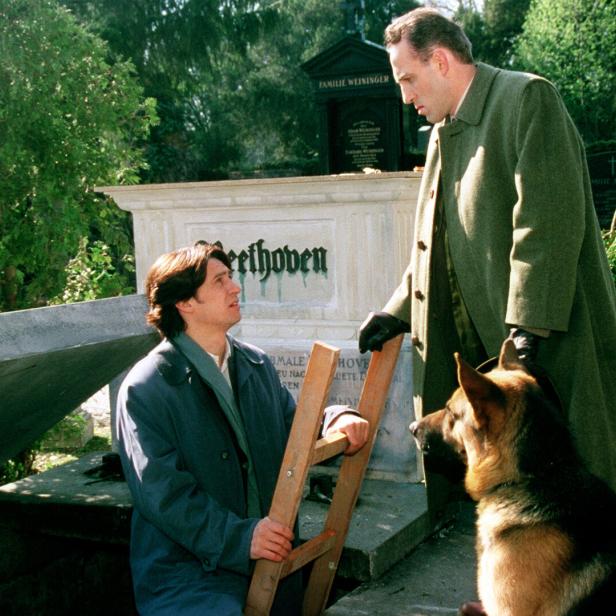 Auf einem Friedhof stehen ein Mann mit Leiter, ein anderer Mann und ein Schäferhund vor einem Grabstein mit der Aufschrift „Beethoven“.