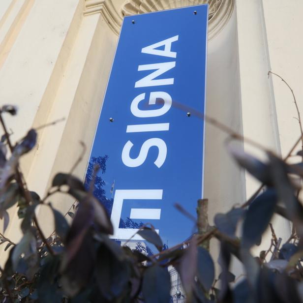 Ein blaues Schild mit der Aufschrift „SIGNA“ an einer Fassade.
