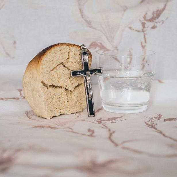 Ein Stück Brot, ein Kreuz und ein Glas Wasser auf einem Tuch mit Blumenmuster.