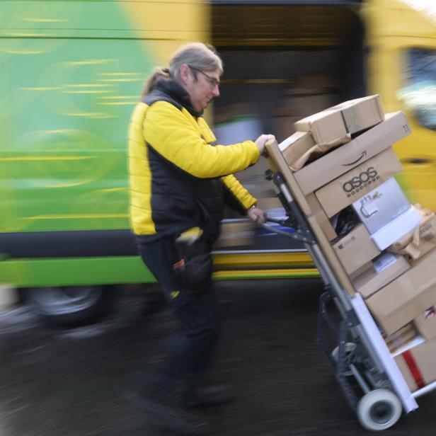 Ein Postbote zieht einen Wagen voller Pakete vor einem Lieferwagen entlang.