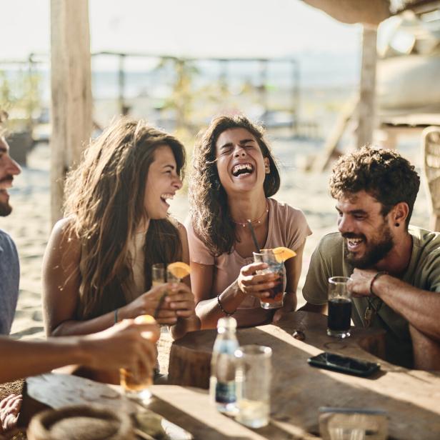 Eine Gruppe junger Leute sitzt an einem Tisch im Freien und lacht, während sie Cocktails trinken.