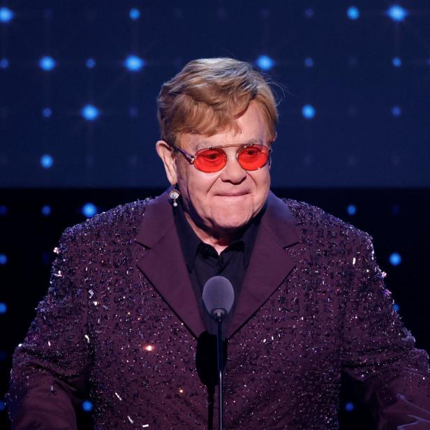 Elton John steht an einem Mikrofon und trägt eine rote Brille.