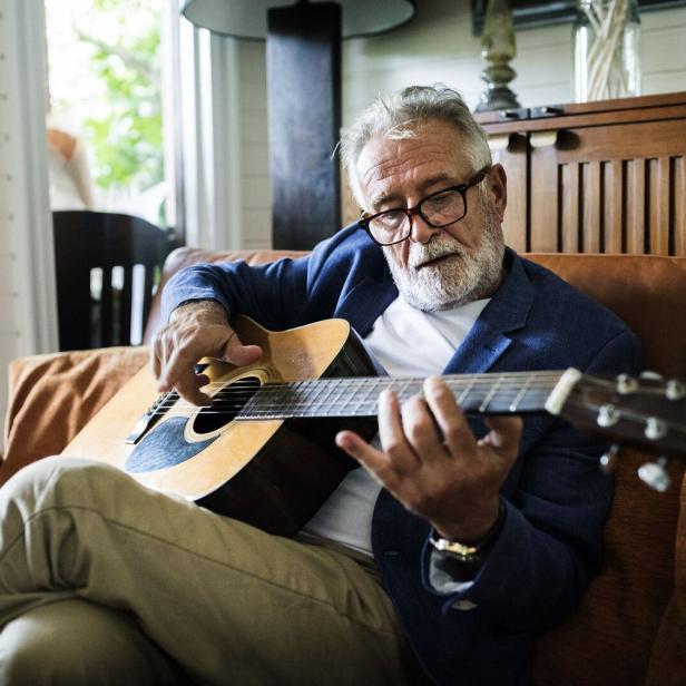Ein älterer Mann mit Brille sitzt auf einem Sofa und spielt Gitarre.