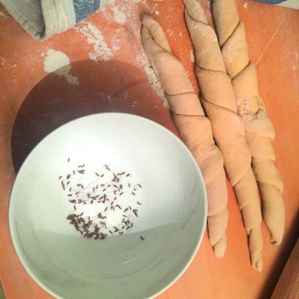 Salzstangerl auf einem Holzbrett, Salz und Kümmel in einer Schüssel neben einer bemehlten Arbeitsfläche.