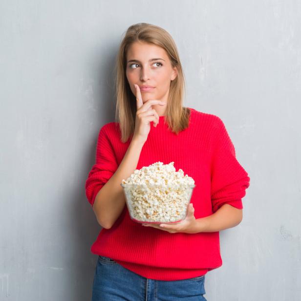 Eine Frau im roten Pullover hält eine Schüssel Popcorn und schaut nachdenklich zur Seite.