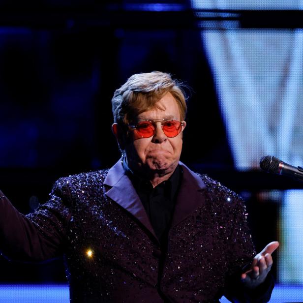 Elton John mit roter Brille und glitzerndem Sakko gestikuliert am Klavier.