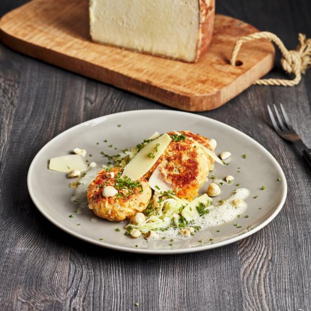 Ein Teller mit Kartoffelpuffern, garniert mit Käse, Kräutern und Haselnüssen, dazu ein Stück Käse auf einem Holzbrett.